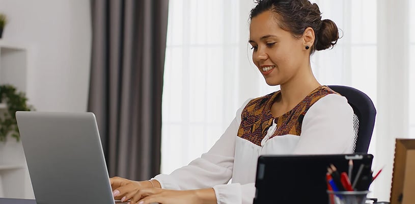 A university student sitting at a desk in her home office, working on a laptop and smiling.